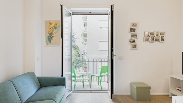 Una sala de estar moderna con un sofá verde, una mesita con dos sillas, un balcón con barandilla y algunas fotos enmarcadas en la pared.