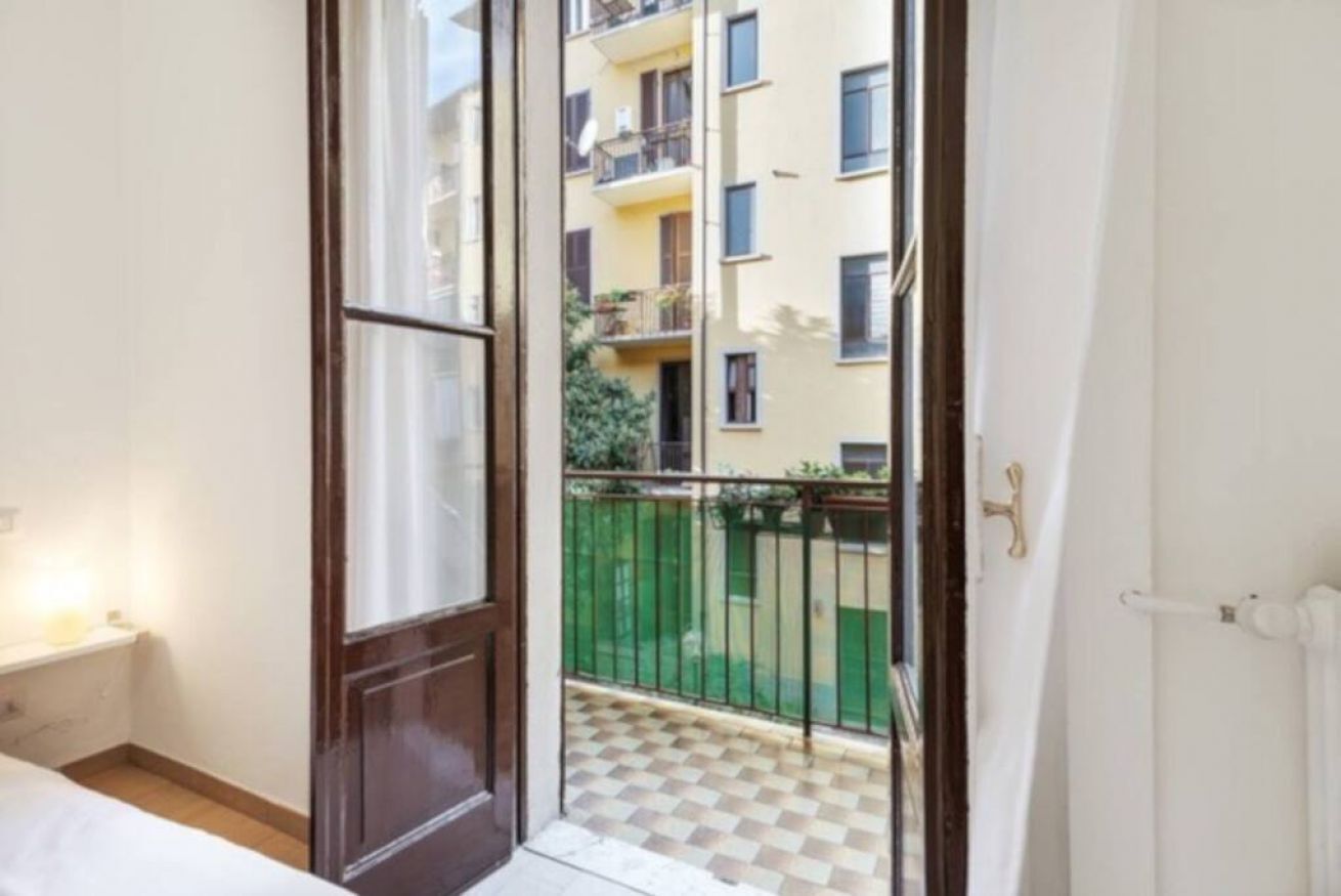 A hotel room with a bed, a small bedside table, a window with a view of a balcony and neighboring buildings.