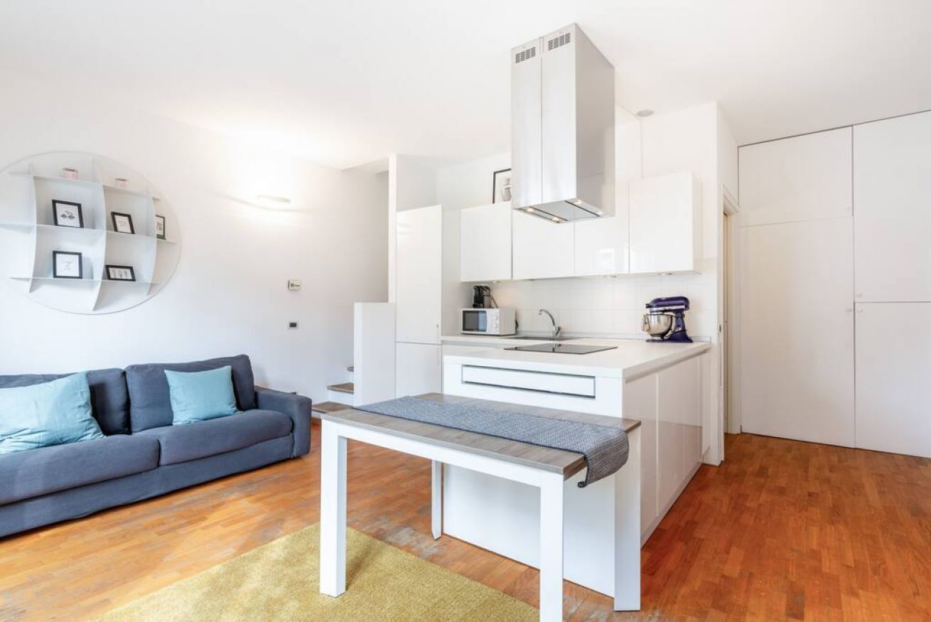 Eine kompakte Küche mit weißen Schränken, einer Mikrowelle und einer Kaffeemaschine. Ein graues Sofa mit blauen Kissen und ein kleiner Tisch mit einer grauen Bank.