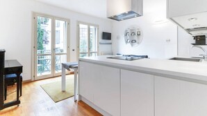 A modern kitchen with white cabinetry, a large island with a sink, and a built-in stove. There is a dining area with a table and chairs, and a view of the outside through large sliding glass doors.