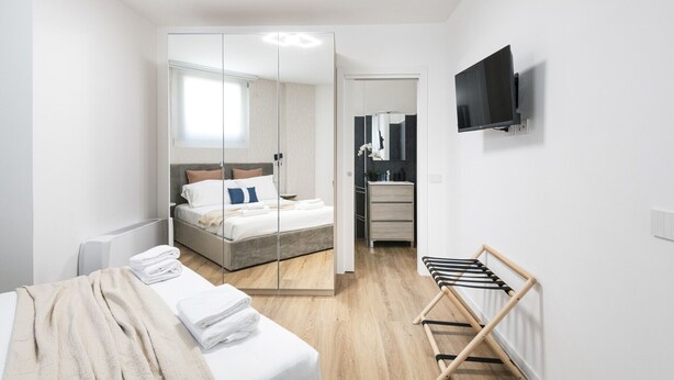 Ein modernes Hotelzimmer mit Bett, Spiegel, Fernseher und einem Wäscheständer aus Holz.