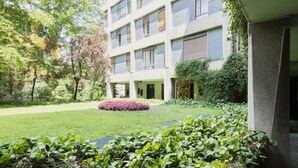 Ein mehrgeschossiges Gebäude mit einem Gartenhof, mit einem Weg, einer kleinen Wasserspiele und üppiger Bepflanzung.