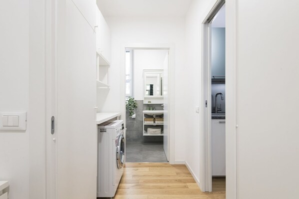 Un pasillo estrecho con una lavadora, estantes y una cocina visible a través de una puerta abierta.