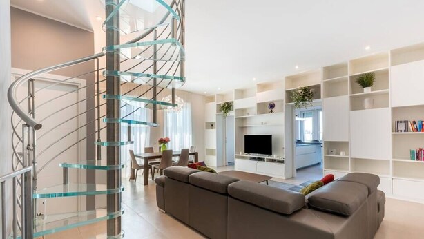Una sala de estar moderna con una escalera de cristal curva, un sofá grande de color gris, un televisor de pantalla plana y estanterías blancas.