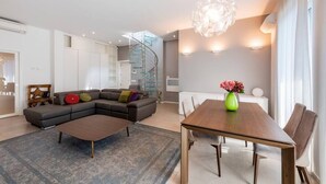 Una sala de estar moderna con un sof á seccional, una mesa de centro de madera y una mesa de comedor con sillas. Se ve una escalera al fondo y un ventanal grande a la derecha.