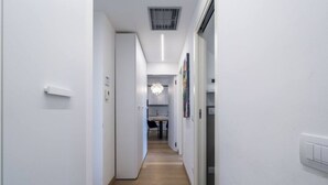 A narrow hallway with white walls and wooden flooring leading to a kitchen area with a chandelier.