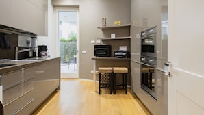 Een moderne keuken met een inbouwoven, magnetron en koffiezetapparaat. Twee barkrukken en een klein plankje met decoratieve spullen.