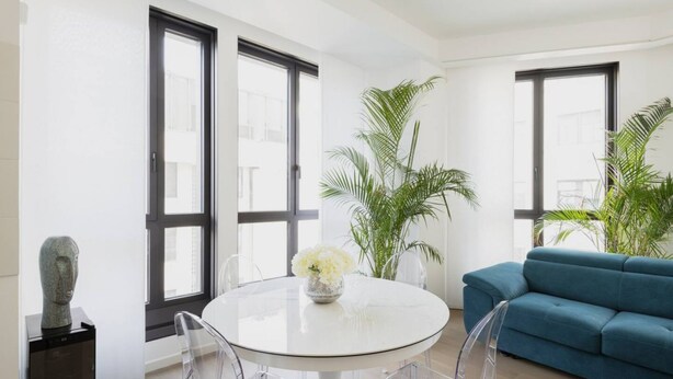 Un salon moderne avec une table ronde blanche, des chaises transparentes, un canapé bleu et de grandes fenêtres.