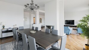 A modern living room with a dark wooden dining table, grey upholstered chairs, a chandelier, a flat-screen TV, and a potted plant.