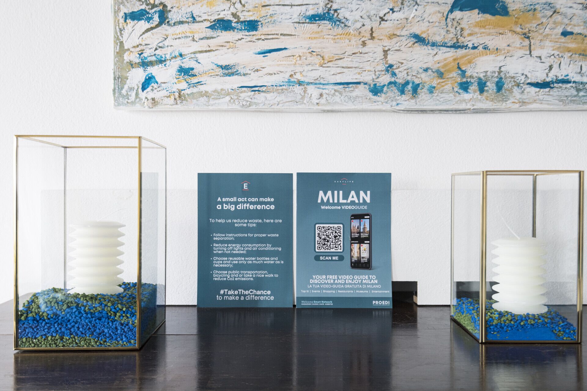 Three glass display cases with white objects and blue pebbles, each with a Milan video guide brochure.