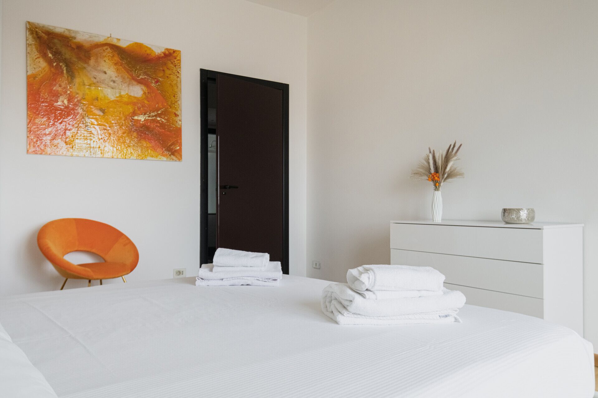 Une chambre d’hôtel moderne avec un grand lit, une commode blanche, un vase contenant des fleurs séchées et un tableau abstrait aux couleurs vives accroché au mur.
