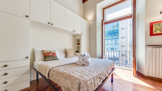 A bedroom with a bed, white wardrobe, and a view of a cityscape through a large window. - Easylife - Refined apartment in Viale Montegrappa, 18 (Milano)