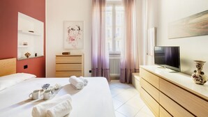 A hotel room with a bed, a TV, a dresser, a window with curtains, and a painting on the wall.