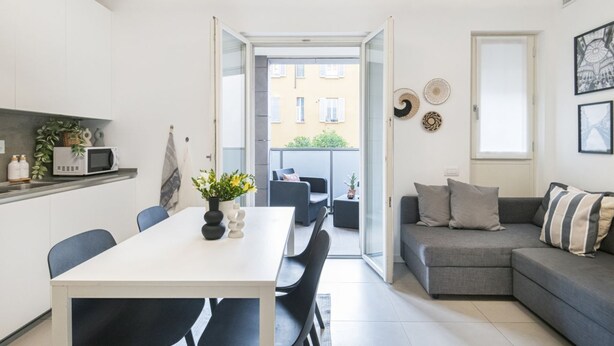 A modern apartment with a dining area, a gray sofa, and a balcony with a view of buildings.