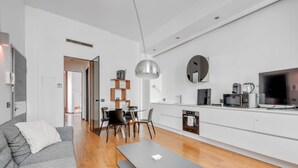 Ein modernes Wohnzimmer mit grauem Sofa, einer Essecke mit Tisch und Stühlen, einer Küche mit weißen Schränken und Geräten sowie einem großen Spiegel an der Wand.