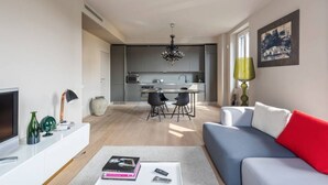 Una sala de estar moderna con televisor, sofá y mesa de centro. Se ve una cocina al fondo, con una zona de comedor.