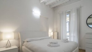 Un dormitorio blanco con cama, lámparas de noche, mesita de noche y cómoda.
