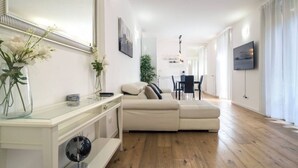 Una sala de estar moderna con un sofá, una mesa de centro de vidrio y un espejo. Hay un televisor montado en la pared y un comedor al fondo.