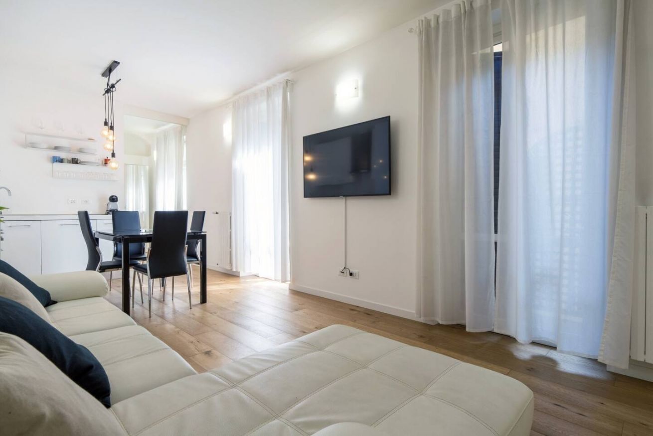 Un soggiorno moderno con divano, zona pranzo con sedie, televisore a parete e un'ampia finestra con tende.