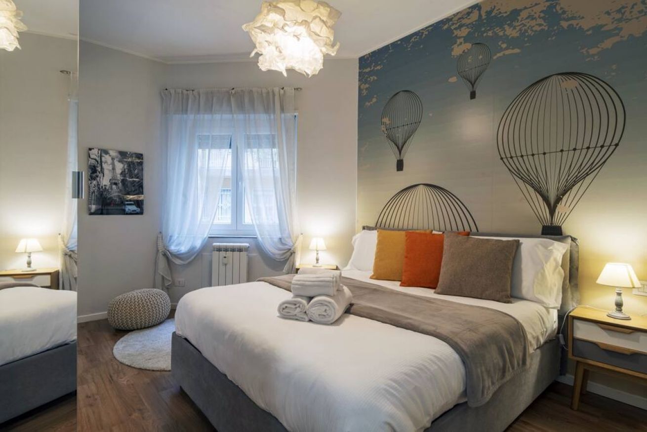 A bedroom with a large bed, two bedside tables with lamps, a round ottoman, a window with sheer curtains, and a wall with hot air balloon artwork.