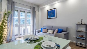 Uma sala de estar moderna com uma mesa de jantar posta para dois, um sofá cinza com almofadas azuis, uma estante preta e uma janela grande com cortinas.