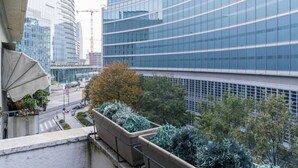 Um edifício moderno com fachada de vidro curva, rodeado por vegetação e uma passarela para pedestres.