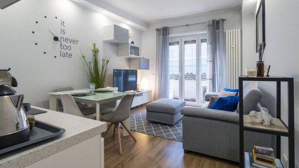 Uma sala de estar moderna com um sofá cinzento, uma área de jantar com mesa e cadeiras, uma cozinha com chaleira e uma janela ampla com cortinas.