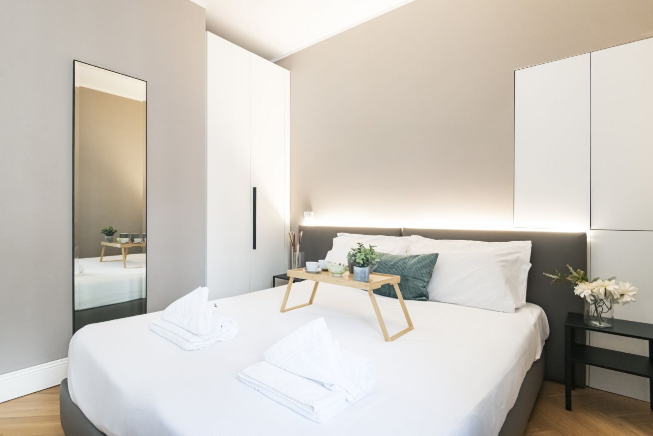 A modern bedroom with a large bed, bedside tables, a mirror, and a nightstand with a vase of flowers.