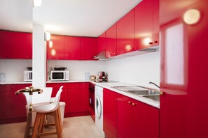 A modern kitchen with red cabinets, a white countertop, a sink, a microwave, a washing machine, and a bar stool.