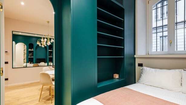 A bedroom with a bed, a window with blinds, a teal-colored wardrobe, and a mirror reflecting a dining area.