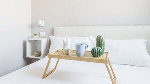 Een net opgemaakt bed met witte lakens, een nachtkastje met een klein plantje en decoratie, en een dienblad met een mok, een klein schaaltje en een cactus.