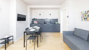 Een moderne keuken met grijze kastjes, een eettafel met stoelen en een grijze bank in een lichte kamer met witte muren.