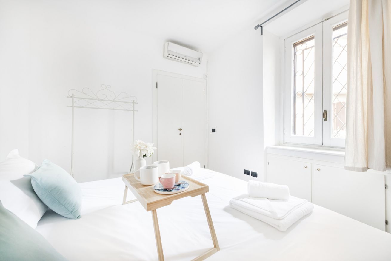 Une chambre d’hôtel lumineuse et blanche, avec un lit, une petite table en bois sur laquelle se trouve un plateau, une fenêtre avec des rideaux et un radiateur.