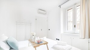 A bright, white hotel room with a bed, a small wooden table with a tray, a window with curtains, and a radiator. - Easylife - Spacious apartment a few steps from Piazza Gae Aulenti (Milan)