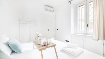 Une chambre d’hôtel lumineuse et blanche, avec un lit, une petite table en bois sur laquelle se trouve un plateau, une fenêtre avec des rideaux et un radiateur.