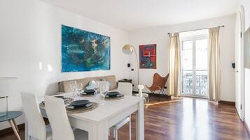 Un coin repas avec une table blanche dressée pour quatre personnes, une grande peinture abstraite accrochée au mur et une vue sur l’extérieur par une porte vitrée.