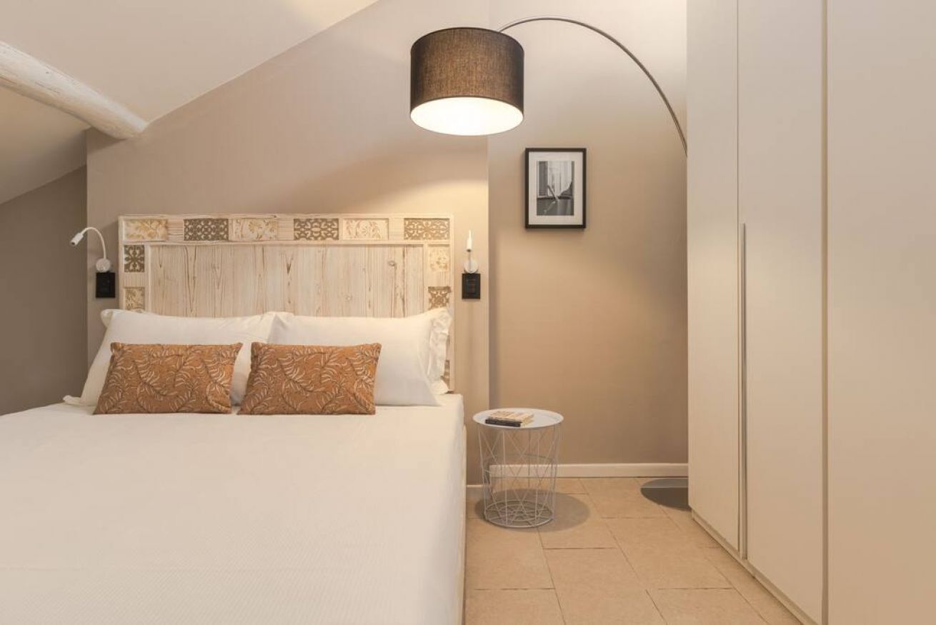 A bedroom with a sloped ceiling, a bed with patterned pillows, a round side table, a wall-mounted lamp, and a framed picture.