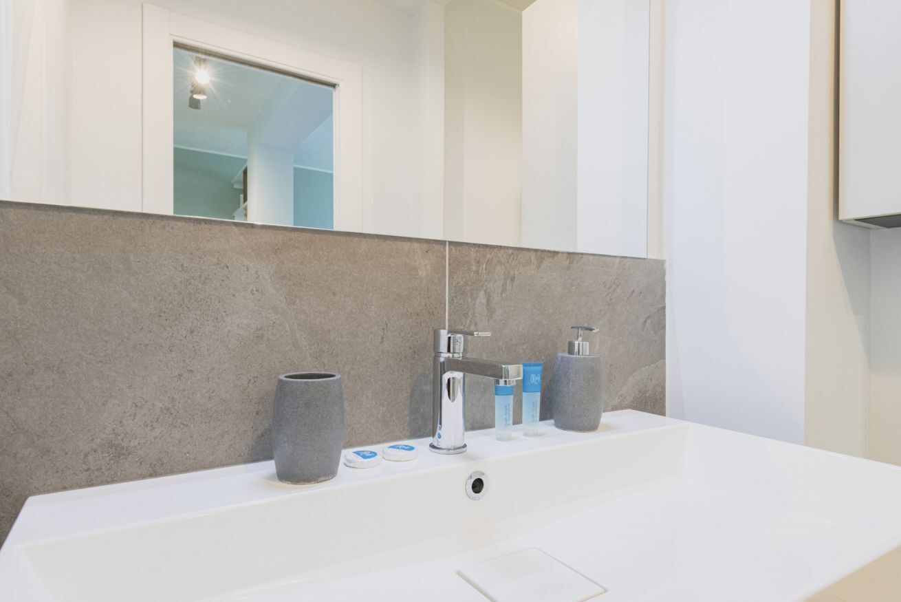 Ein modernes Badezimmer mit einem weißen Waschbecken, einer grauen Steinwand und einem Fenster über dem Spiegel.
