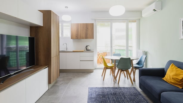 Una cocina moderna con armarios blancos, detalles de madera, un televisor integrado y un comedor con sillas amarillas.