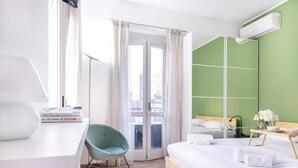 A modern hotel room with a wooden bed, a desk with books, a green chair, and a view through a glass door.