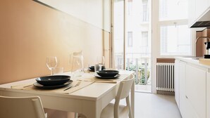A modern dining area with a table set for two, a window with a view of the city, and a kitchen area visible through the open door. - Easylife - Bright residence in Sant'Ambrogio (Milano)