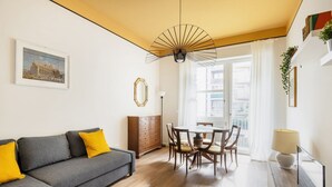 A modern living room with a gray sofa, yellow cushions, a wooden dining table with chairs, a ceiling fan, and a large window with curtains. - Easylife - Bright residence in Sant'Ambrogio (Milano)