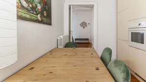 A narrow room with a wooden table and green chairs, a radiator, a bench, and a white cabinet with an oven.