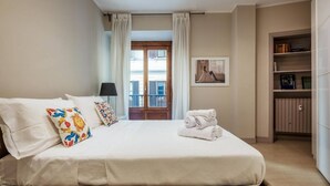 Habitación de hotel con una cama grande, una mesita de noche, una estantería y vistas al exterior a través de una ventana con cortinas.