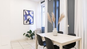 Un espace repas moderne avec une table blanche, des chaises noires, un vase contenant des plantes séchées et un tableau abstrait bleu accroché au mur.