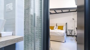 Una habitación de hotel moderna con cabina de ducha de vidrio, una mesita de noche con un objeto blanco, una cama con ropa de cama blanca y un almohadón amarillo, y una pequeña mesa con una lámpara.