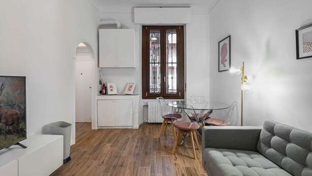 A modern living room with a grey sofa, a dining area with a glass table and chairs, and a kitchen with white cabinets and a radiator. - Easylife - Cuddle & relax in the heart of Isola (Milano)
