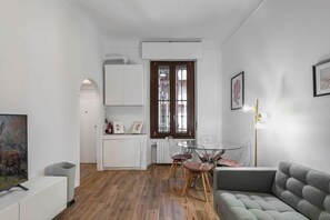 A modern living room with a grey sofa, a dining area with a glass table and chairs, and a kitchen with white cabinets and a radiator.