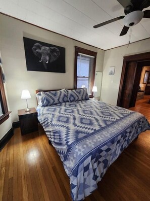A bedroom with a patterned bedspread, two bedside lamps, a ceiling fan, and a framed elephant artwork on the wall.