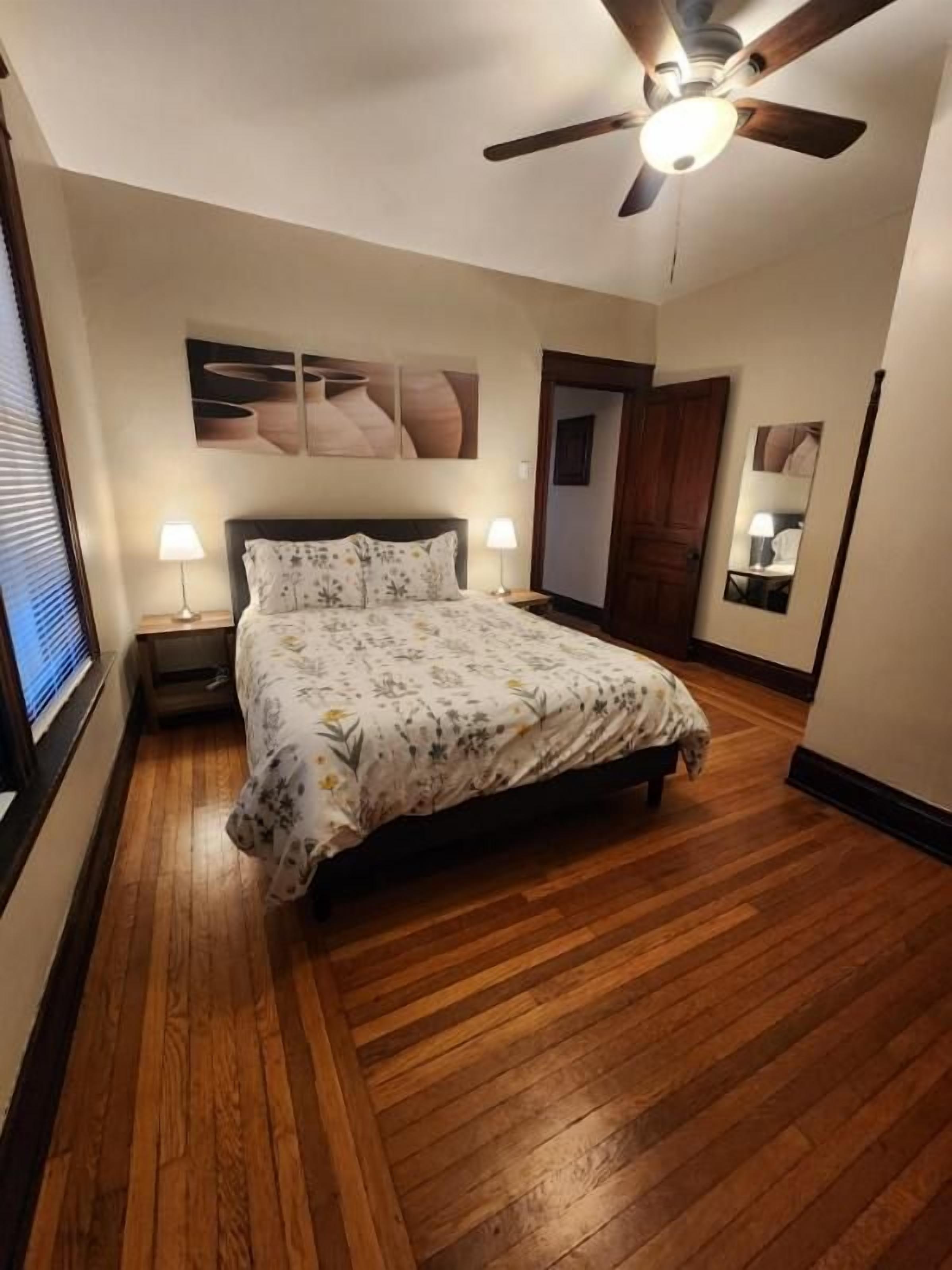 Un dormitorio con piso de madera, una cama con colcha floral, dos lámparas de noche, un ventilador de techo y una obra de arte colgada en la pared.